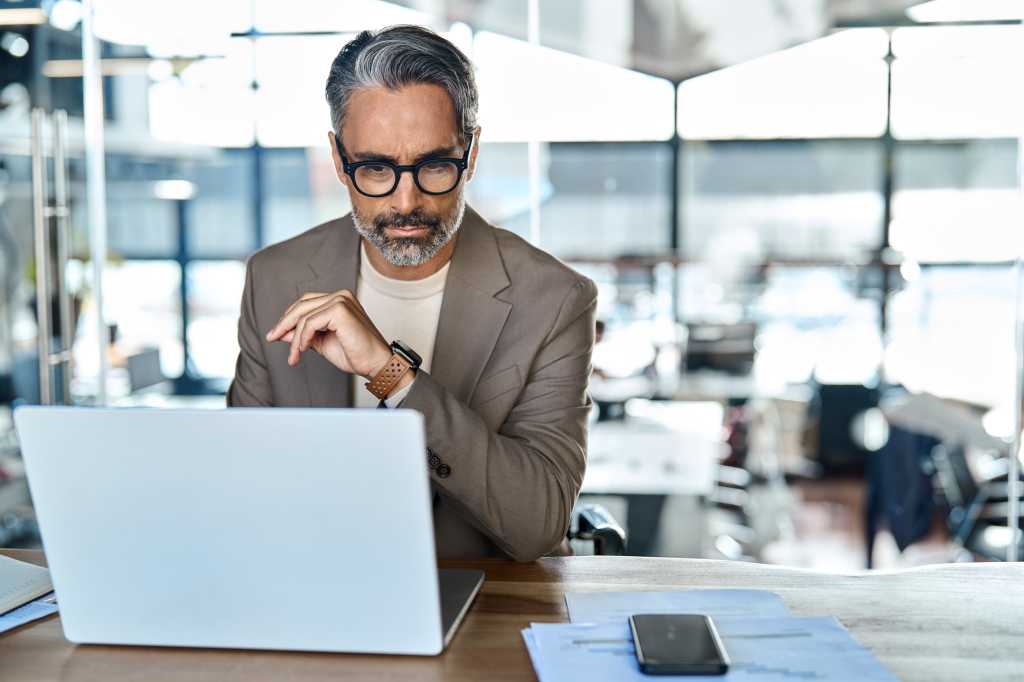 Busy serious mature business man executive sitting at desk using laptop. Professional middle aged businessman ceo lawyer investor working on computer thinking on investment question in office.