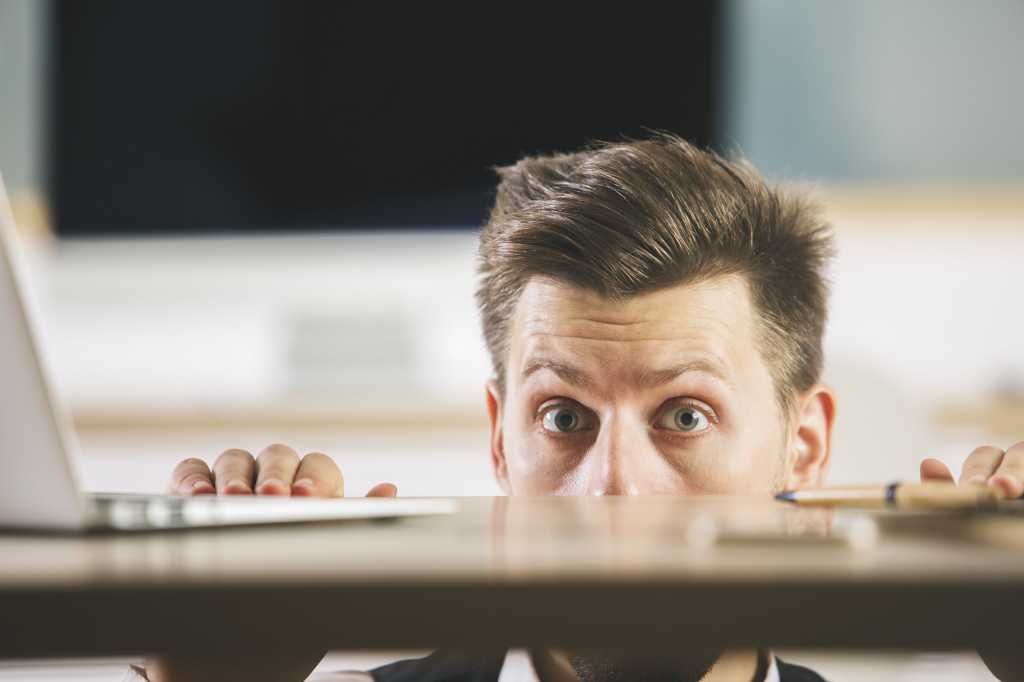 Man hiding behind blurry wooden desktop with laptop. Fearful anxious worker.